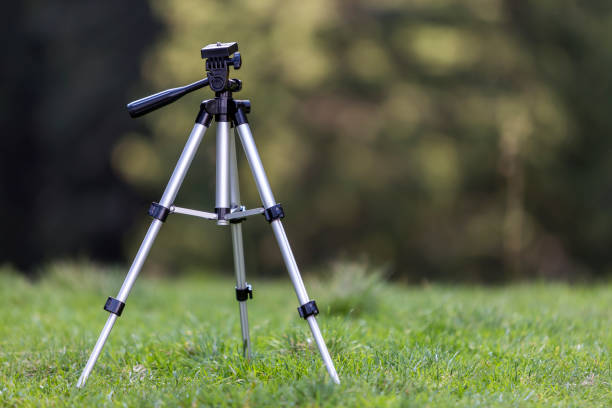 istock Isolated aluminum shiny tripod standing alone in fresh grassy meadow on bright sunny summer day on blurred dark green forest trees foliage bokeh copy space background. Photography as job and hobby. 1068416174