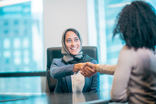 A woman wearing a head scarf is shaking hands with another woman after making a deal. They are indoors in an office building.