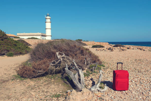 istock Lighthouse and red suitcase on a rocky shore at Cap de Ses Salines. Selective focus 1068423312