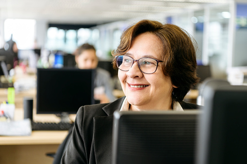 Cheerful businesswoman in her 50s smiling and looking away, using pc, sitting at her desk