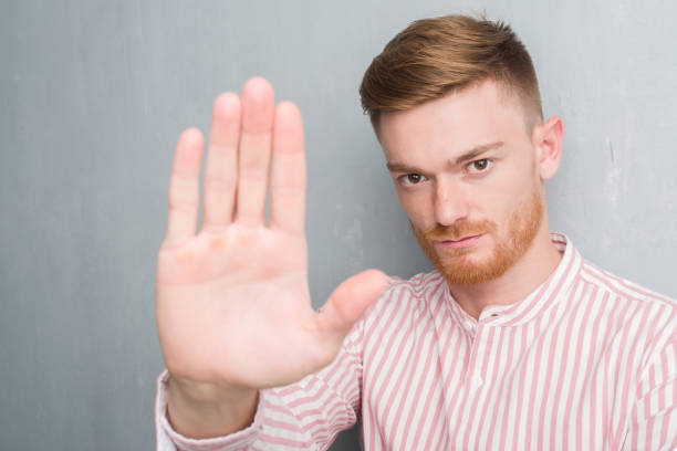 istock Young redhead man over grey grunge wall with open hand doing stop sign with serious and confident expression, defense gesture 1068445896