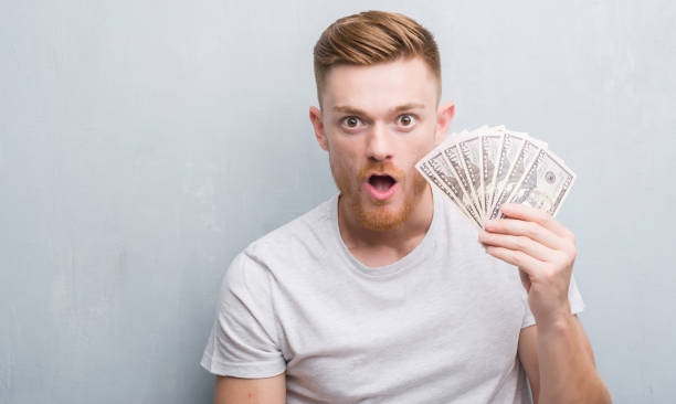 istock Young redhead man over grey grunge wall holding dollars scared in shock with a surprise face, afraid and excited with fear expression 1068446596