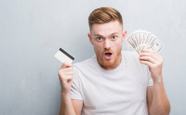 istock Young redhead man holding dollars and credit card scared in shock with a surprise face, afraid and excited with fear expression 1068446672