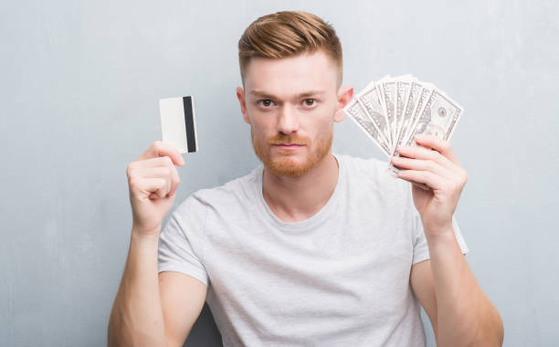 istock Young redhead man holding dollars and credit card with serious expression on face. Simple and natural looking at the camera. 1068446848