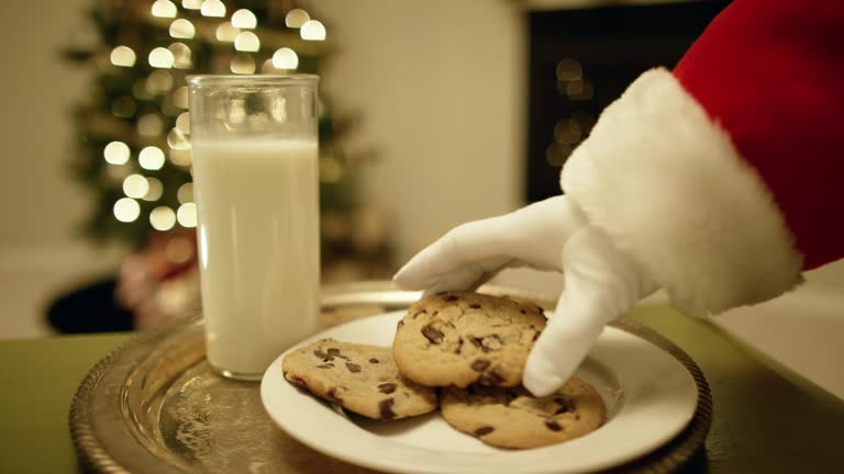 Santa Claus' Gloved Hand Picks Up a Chocolate Chip Cookie from a Tray with a Glass of Milk on It with a Christmas Tree and a Fireplace in the Background on Christmas Eve