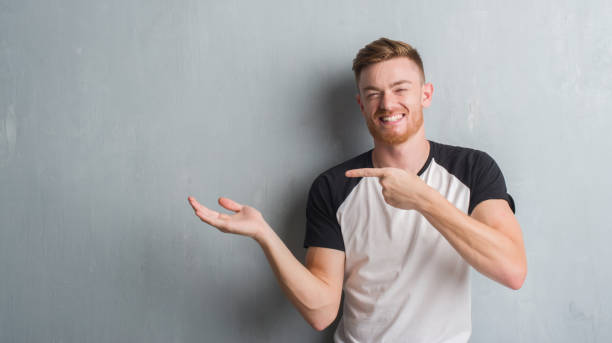 istock Young redhead man over grey grunge wall amazed and smiling to the camera while presenting with hand and pointing with finger. 1068448940