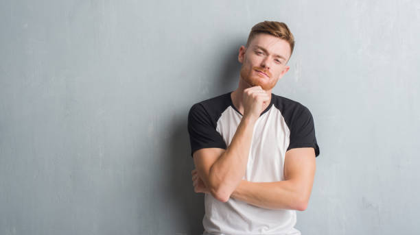 istock Young redhead man over grey grunge wall looking confident at the camera with smile with crossed arms and hand raised on chin. Thinking positive. 1068448988