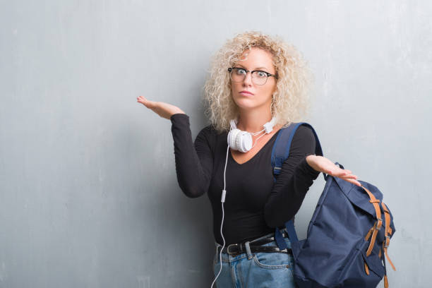 young blonde woman over grunge grey wall wearing backpack and headphones clueless and confused expression with arms and hands raised. doubt concept. - head and shoulders audio imagens e fotografias de stock