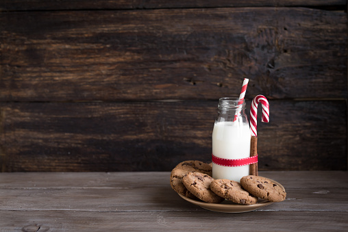 Cookies and Milk for Santa. Traditional Christmas homemade chocolate chip cookies, candy cane and bottle of milk with red ribbon on rustic wooden table, copy space.