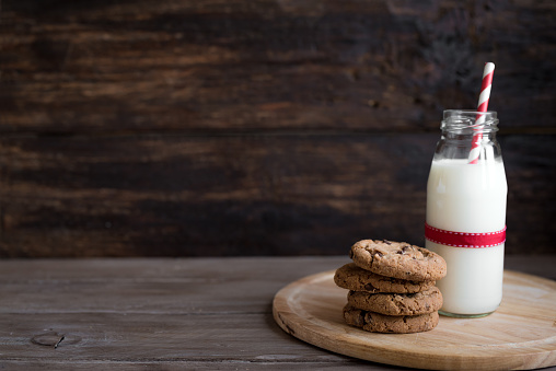 Cookies and Milk for Santa. Traditional Christmas homemade chocolate chip cookies and bottle of milk with red ribbon on rustic wooden table, copy space.