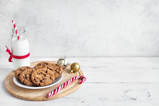 Chocolate chip cookies and Milk for Santa. Traditional Christmas homemade cookies, candy cane and bottle of milk with Christmas decor on white wooden table, copy space.