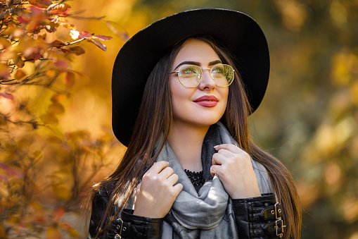 Close-up portrait of cheerful smiling woman in glasses, black dress and hat on background of yellow autumn leaves. fashionable girl