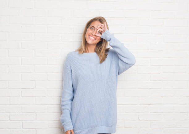 istock Beautiful young woman over white brick wall doing ok gesture with hand smiling, eye looking through fingers with happy face. 1068467368