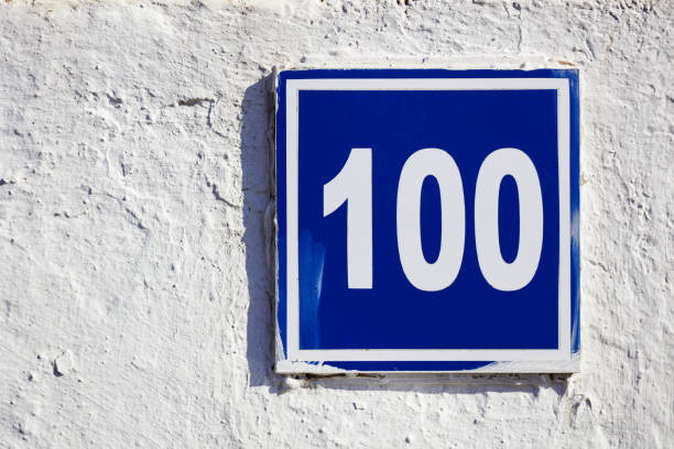 Outdoor house number one hundred 100 on blue square metal plate mounted on a white textured wall