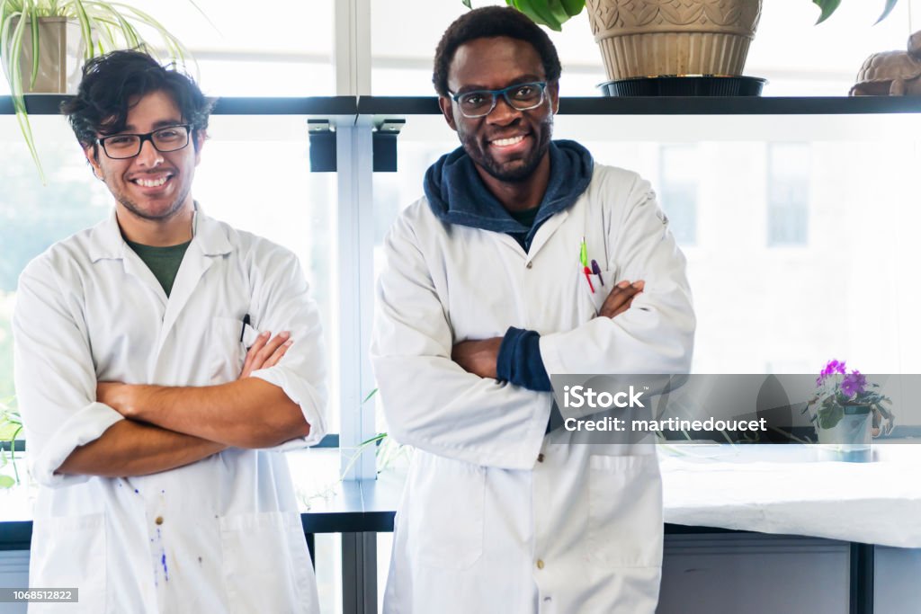 Multi-ethnic duo of professors in college science laboratory. Multi-ethnic duo of professors in college science laboratory. They are looking at the camera, with a big smile and arms crossed. They are wearing lab coats. Horizontal waist up large view indoors shot with copy space. Occupation Stock Photo