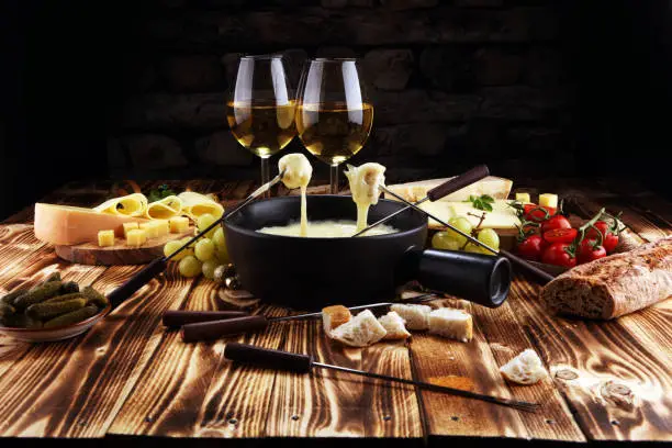 Gourmet Swiss fondue dinner on a winter evening with assorted cheeses on a board alongside a heated pot of cheese fondue with two forks dipping bread and white wine behind in a tavern or restaurant