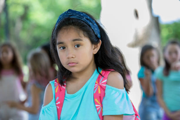 istock Sad Asian elementary schoolgirl looks into camera while classmates in background bully her 1068597326