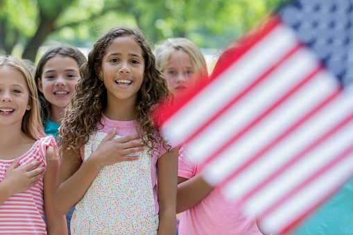 Biracial African American and Caucasian elementary age little girl is standing outdoors with diverse classmates. Students are reciting Pledge of Allegiance to the American Flag.