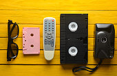 Audio cassette, vhs, 3d glasses, tv remote, hipster film camera on a yellow wooden background. Retro devices from 80s. Top view.
