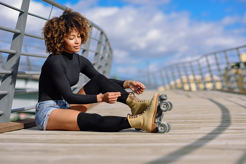Young smiling black girl sitting on urban bridge and puts on skates. Woman with afro hairstyle rollerblading on sunny day