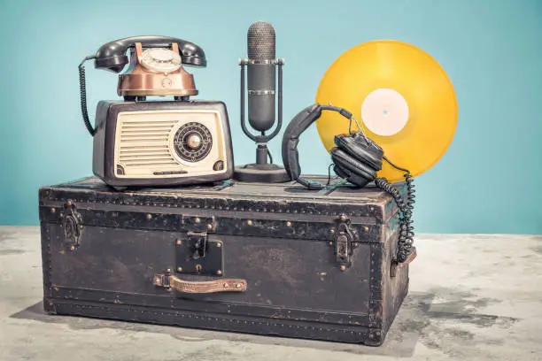 Photo of Retro radio from 60s, old copper telephone, studio microphone from 50s, and gold colored vinyl disk record circa 70s, headphones on aged classic travel trunk. Vintage style filtered photo