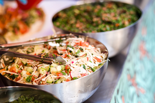 Close up color image depicting freshly prepared salads suitable for vegetarians and vegans for sale at an outdoor food market in London, UK. The salads consist of onion, cucumber, tomatoes and a variety of herbs and dressings. They are presented in metal bowls in a row, with diminishing perspective. Room for copy space.