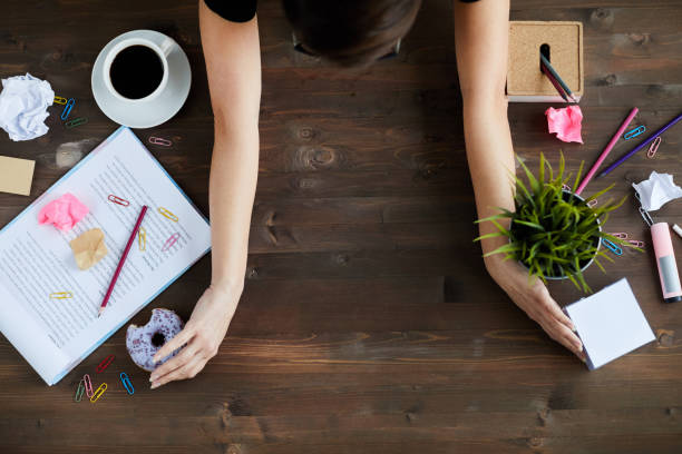 Woman Cleaning Up Office Table Top view of unrecognizable woman clearing up space on wooden desk, putting away stationery, papers, plant, food and other stuff messy vs clean desk stock pictures, royalty-free photos & images