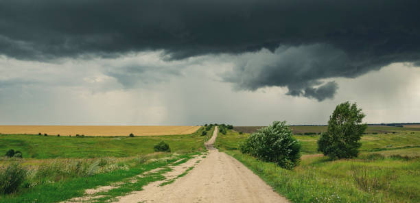 istock Cloudy summer landscape with ground country road passing through the fields on a background of dark overcast sky. 1068852150