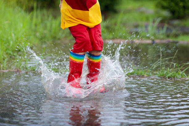 Kid playing out in the rain. Children with umbrella and rain boots play outdoors in heavy rain. Little boy jumping in muddy puddle. Kids fun by rainy autumn weather. Child running in tropical storm.