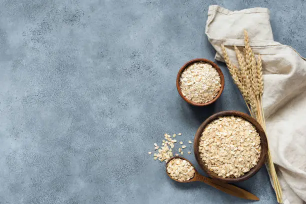 Rolled oats, oat flakes, whole grain oats in wooden bowl, linen textile and golden ears of wheat on concrete background with copy space for text