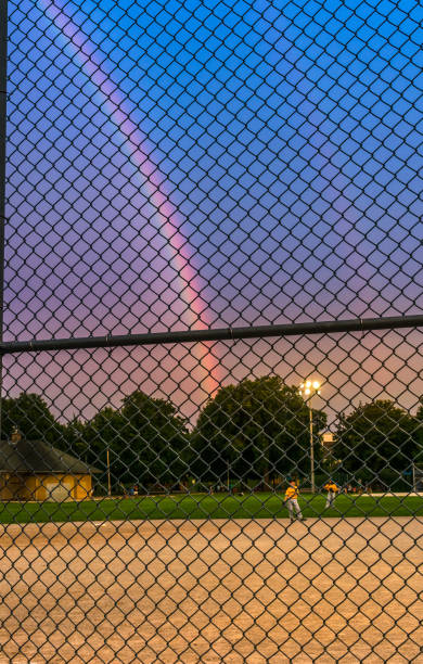 View from sidelines, at little league baseball game, at dusk, unidentifiable players out of focus, chain link fence are focus. Rainbow twilight sky.