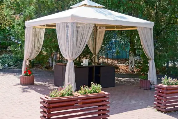 open gazebo with curtains and furniture on the sidewalk in the park among green trees