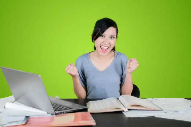 Pretty college student celebrating her success with books and laptop computer on the table