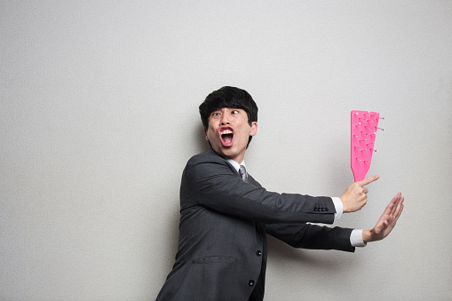 Japanese businessman who has japanese new year ornament.
He wearing gray suit, and stand in front of white wall.
He has a pink object what designed in the motif of Japanese new year ornament.