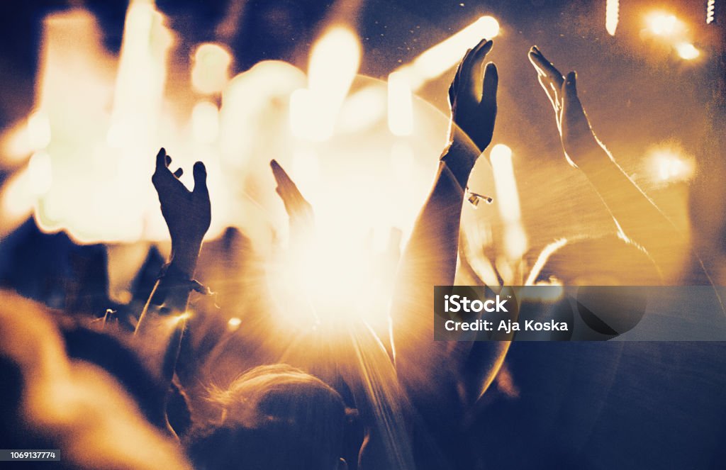 Cheering fans at concert. Rear view of large group of unrecognizable people at concert. Their hands are in the air, clapping. Beige stage lights in the background. Two people in foreground are released. Party - Social Event Stock Photo