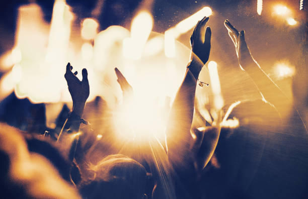 Cheering fans at concert. Rear view of large group of unrecognizable people at concert. Their hands are in the air, clapping. Beige stage lights in the background. Two people in foreground are released. nightlife photos stock pictures, royalty-free photos & images