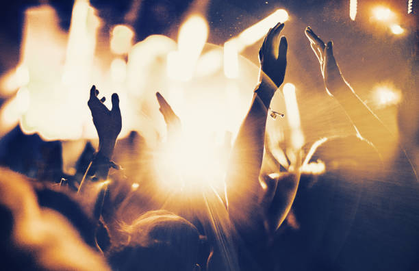 Cheering fans at concert. Rear view of large group of unrecognizable people at concert. Their hands are in the air, clapping. Beige stage lights in the background. Two people in foreground are released. Applauding stock pictures, royalty-free photos & images