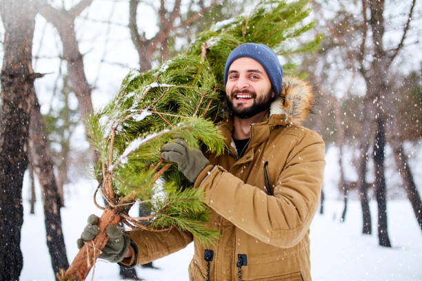 istock Bearded man carrying freshly cut down christmas tree in forest. Young lumberjack bears fir tree on his shoulder in the woods. Irresponsible behavior towards nature, save forest, keep green concept. 1069143868