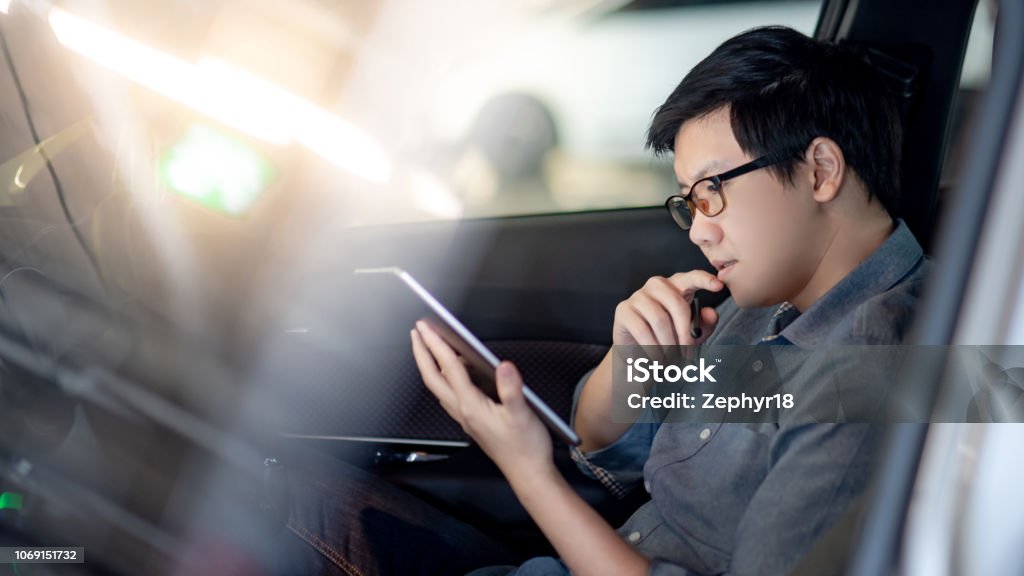 Young Asian businessman with glasses reading news on digital tablet while sitting on driver seat in his car. Business and technology concept Asian and Indian Ethnicities Stock Photo