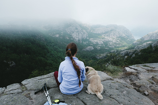 Woman with long hair and small pug sit at the edge of the cliff looking and beautiful mountains in South Norway