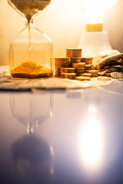 istock Sand running through the shape of hourglass on table with banknotes and coins of international currency. Time investment and retirement saving. Urgency countdown timer for business deadline concept 1069154372
