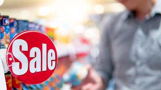 Red sale label on product shelf in supermarket with blurred male shopper choosing food package in the background. shopping lifestyle in grocery store concept