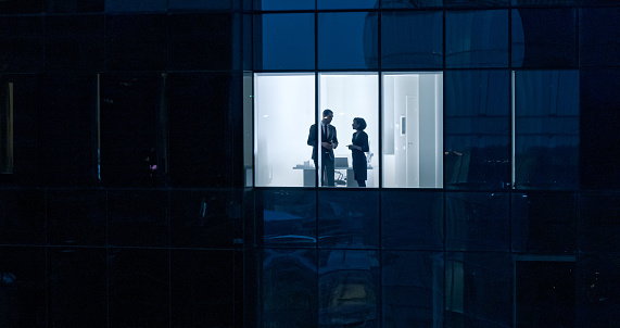 Aerial Shot From Outside of the Skyscraper: Businessman and Businesswoman Talking while Standing in the Office Window. Shot of the Business District in the Evening and Businesspeople Working in the Big City.