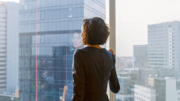 photo de la femme d’affaires dans une robe élégante dans son bureau à la recherche hors de la fenêtre de manière réfléchie. bureau d’affaires moderne avec des ordinateurs personnels et vue sur la grande ville. - back people rear view looking at view photos et images de collection