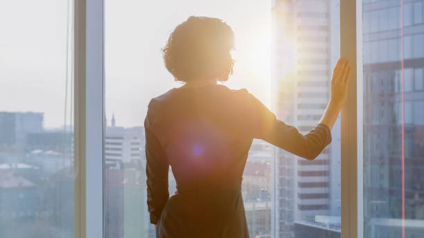 photo de la femme d’affaires dans une robe noire frappant dans son bureau regarde par la fenêtre au coucher du soleil. bureau d’affaires moderne avec des ordinateurs personnels et vue sur la grande ville. - regarder par la fenêtre photos et images de collection