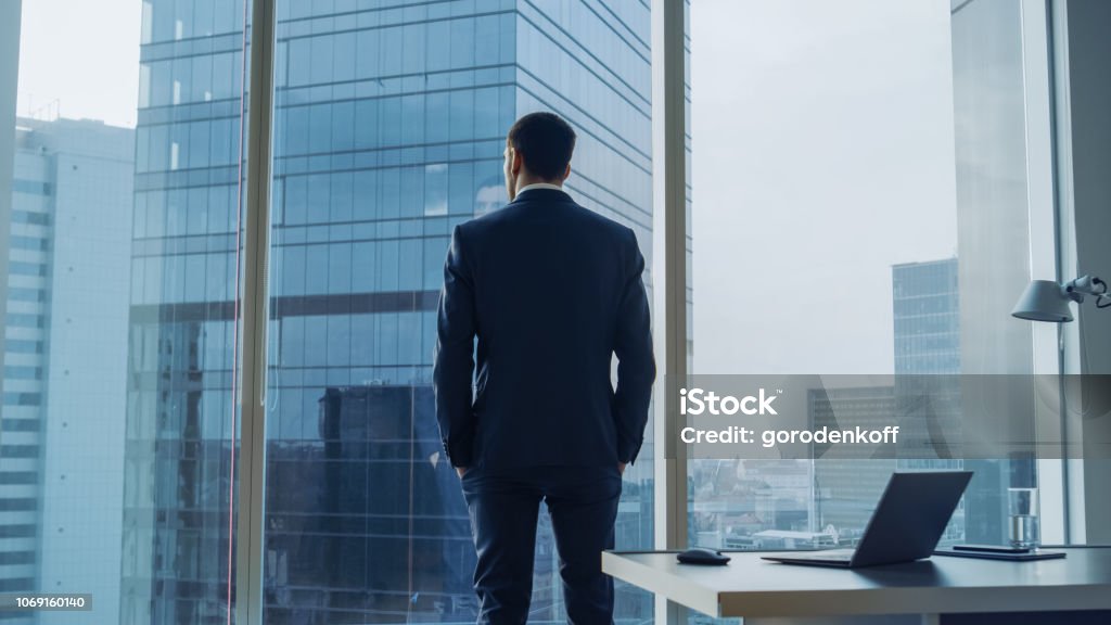 Vue de l’homme d’affaires pensif, vêtu d’un costume debout dans son bureau, mains dans les poches et contemplant Next Big Business Deal, regarde par la fenêtre de l’arrière. Vue de fenêtre panoramique grand quartier des affaires. - Photo de Homme d'affaires libre de droits