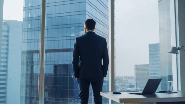 vue de l’homme d’affaires pensif, vêtu d’un costume debout dans son bureau, mains dans les poches et contemplant next big business deal, regarde par la fenêtre de l’arrière. vue de fenêtre panoramique grand quartier des affaires. - winning success business businessman photos et images de collection