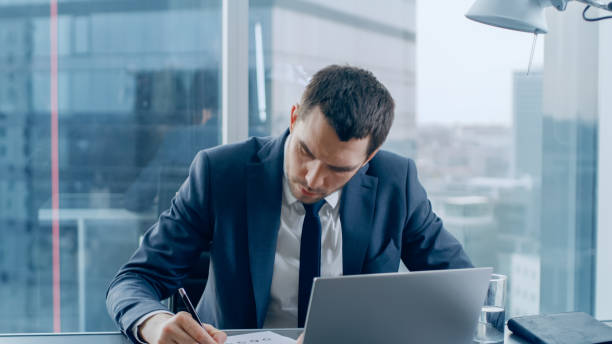 istock Successful Businessman Sitting at His Desk, Working with Papers, Signing Documents and Using Laptop. In the Window Big City Business District View. 1069160208