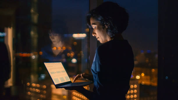 empresaria exitosa, elegante vestido tiene laptop estando cerca de la ventana de su oficina. tarde en la noche mujer profesional haciendo trabajo importante. la ventana tiene vista distrito de negocios de ciudad grande con muchas luces de la noche. - night cityscape reflection usa fotografías e imágenes de stock