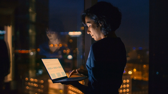 Successful, Stylishly Dressed Businesswoman Holds Laptop While Standing Near the Window of Her Office. Late at Night Professional Woman Doing Important Job. Window Has Big City Business District View with Many Night Lights.